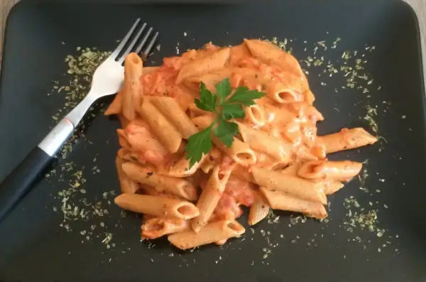 Penne w kremowym sosie pomidorowym z mascarpone, udekorowane natką pietruszki. Obok widelec i posypka z ziół.
