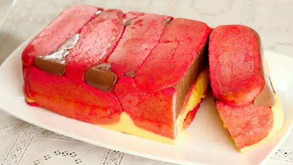 Una fetta di zuppa inglese tradizionale, con strati di pan di Spagna rosa, crema pasticcera gialla e cioccolato, decorata con savoiardi rossi e gocce di cioccolato.