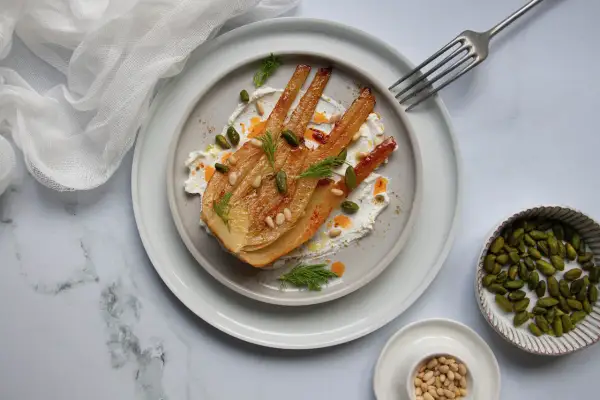 Un plat de fenouil au four, doré et tendre, servi sur une crème onctueuse, garni de pistaches, pignons et d'aneth frais.