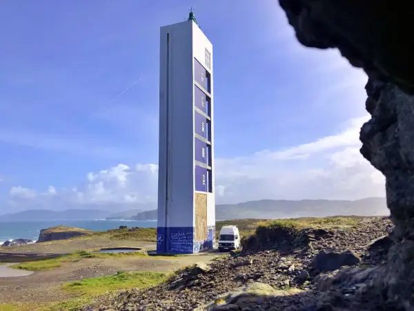 El faro de Punta Frouxeira se alza imponente sobre la costa, con un cielo azul y nubes blancas. Una furgoneta blanca descansa a su lado.