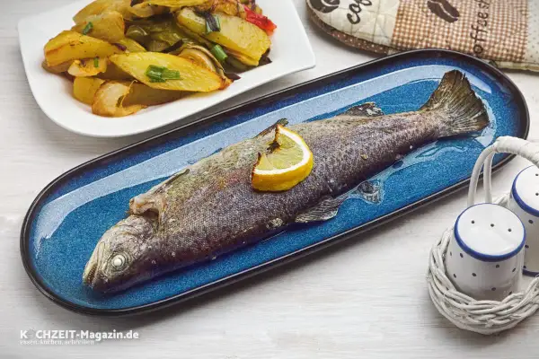 Ein ganzer, im Ofen gebackener Fisch auf einem blauen Teller, garniert mit einer Zitronenspalte. Daneben eine Portion Kartoffel-Gemüse-Spalten.