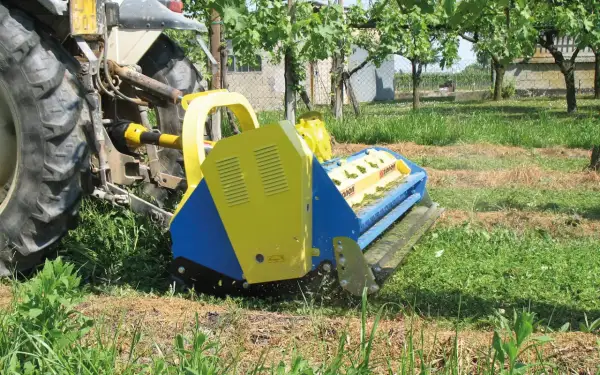 Trattore con falciatrice gialla e blu che lavora in un vigneto. L'erba tagliata copre il terreno, un segno di potenziale inquinamento del suolo se non gestita correttamente.