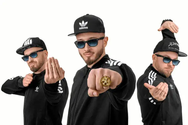 Trzech mężczyzn w czarnych bluzach z paskami Adidas i czapkach z daszkiem, pozujących jak zespoły disco polo.
