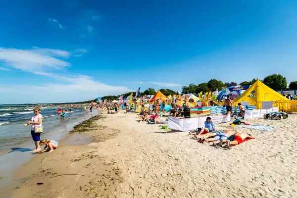 Słoneczna plaża w Ustce pełna ludzi, kolorowych namiotów i flag. Wiele atrakcji dla każdego.