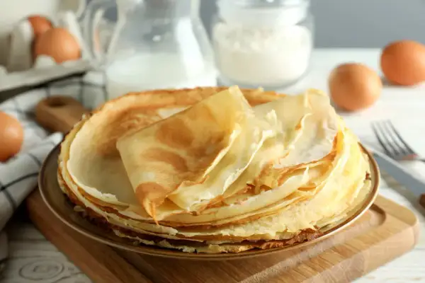 Pila di crepes dorate, pronte per essere farcite. Ingredienti per una ricetta crepes dolci perfetta: uova, latte e farina sullo sfondo.