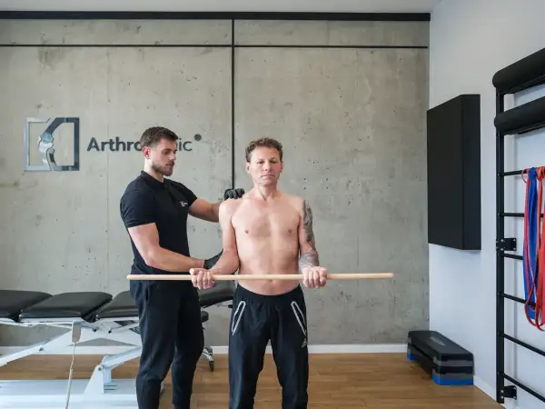 Fizjoterapeuta pomaga pacjentowi w ćwiczeniach rehabilitacja barku po operacji. Mężczyzna trzyma kij, wykonując ruchy ramionami.