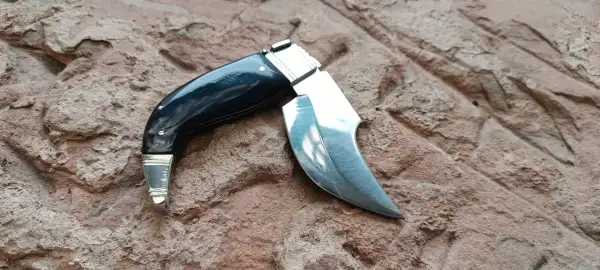 Navaja capaora de mango negro y hoja curva, lista para la acción sobre una superficie rocosa.