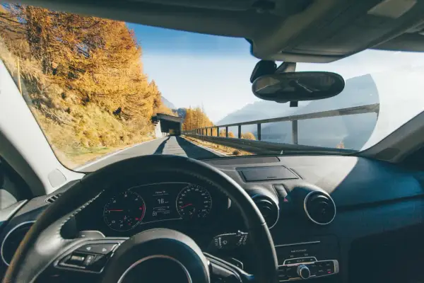 Volant d'une voiture en mouvement, tableau de bord affichant 16 km/h. Paysage d'automne en Italie, arbres dorés, montagnes et tunnel.