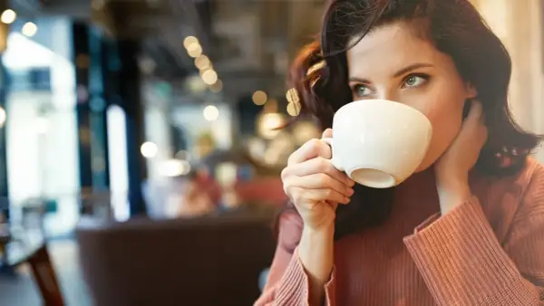 En kvinna med mörkt hår dricker kaffe ur en vit kopp. Hon har en stickad tröja och sitter på ett café. Kanske lite kaffe på tom mage för att vakna till.