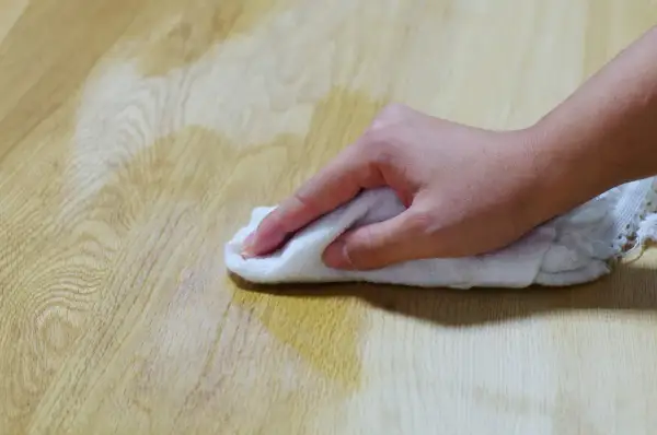 Main d'une personne essuyant une tache d'huile sur un parquet en bois avec un chiffon blanc.
