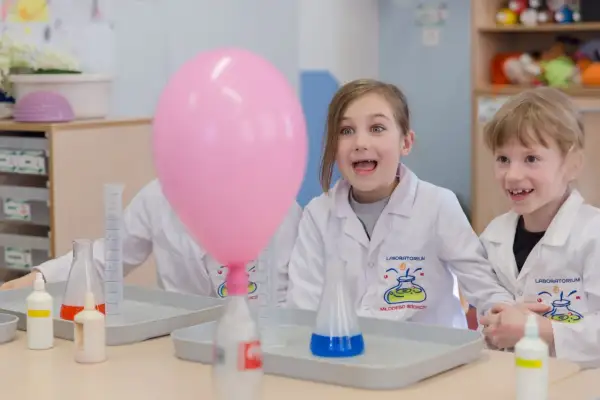 Dzieci w fartuchach laboratoryjnych z ekscytacją obserwują różowy balon podczas eksperymentów chemicznych dla dzieci.