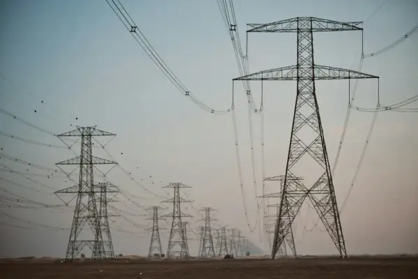Linia wysokiego napięcia z wieżami energetycznymi, przypominającymi gigantyczne kogeneratory, ciągnie się przez pustynny krajobraz.