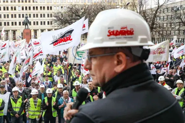 Mężczyzna w białym kasku z logo "Solidarność" przemawia do tłumu ludzi z flagami. To demonstracja związków zawodowych.