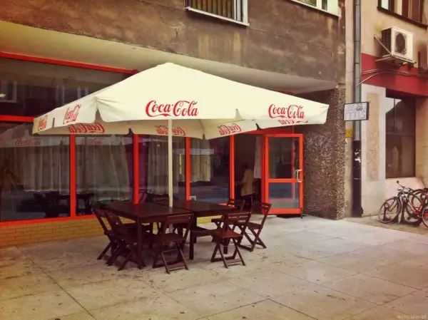 Tanie jedzenie Wrocław Rynek: stoliki pod parasolem Coca-Cola, wejście do lokalu, rowery przy ścianie.