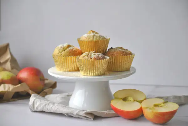 Goldene Muffins mit Puderzucker, daneben Äpfel und eine Tüte. Ein leckeres apfelmuffins rezept zum Nachbacken.