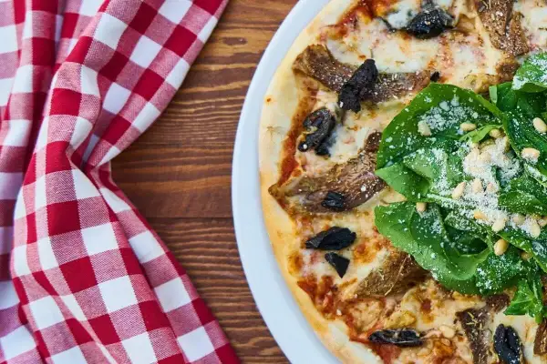 Pizza z rukolą i grzybami na białym talerzu, obok czerwono-biała kraciasta serwetka.