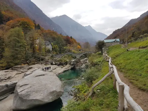 Sentiero sul monte Valdo, con un ponte ad arco in pietra, un fiume turchese e montagne sullo sfondo.