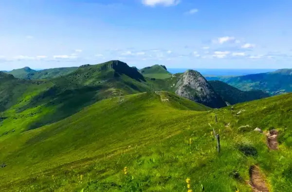 Paysage de montagnes verdoyantes sous un ciel bleu, idéal pour les plus belles randonnées dans le Cantal. Un sentier serpente sur la colline.