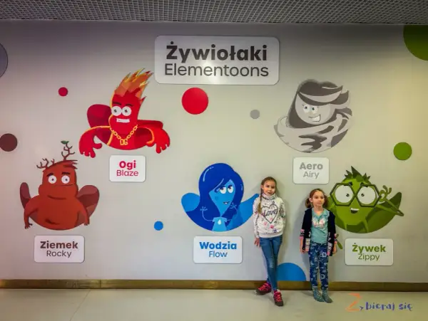 Dwie dziewczynki pozują na tle ściany z postaciami z bajki "Żywiołaki Elementoons". Widać Ogi Blaze, Ziemka Rocky, Wodzię Flow, Aero Airy i Żywka Zippy.
