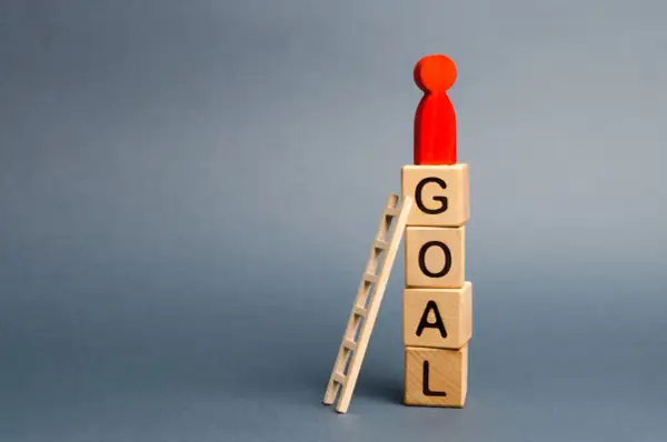 Drewniane klocki z literami GOAL, drabinka i czerwona figurka symbolizująca cel.