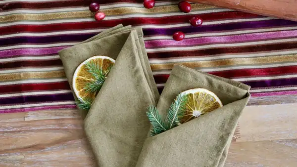 Un pliage serviette original avec une tranche d'orange séchée et une branche de sapin, sur une nappe rayée aux couleurs vives.