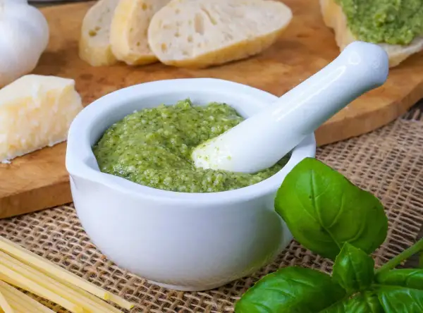 Frisches Pesto in einem Mörser, bereit zum Haltbarmachen mit Salz. Daneben liegen Brot, Parmesan, Knoblauch und Pasta.