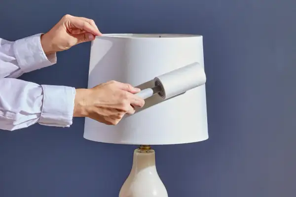 Hands using a lint roller to clean a white lampshade, demonstrating how to clean lamp shades effectively.
