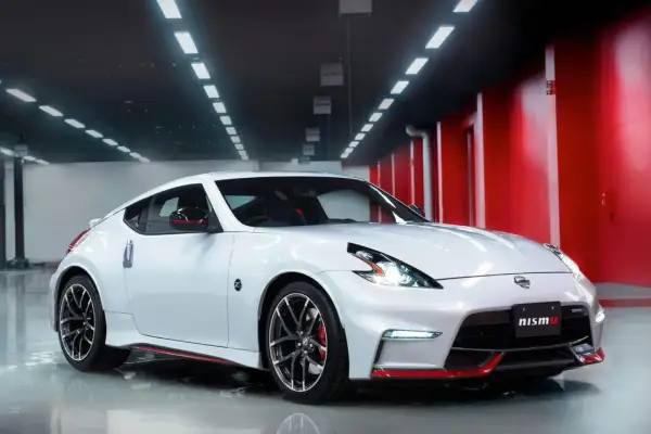 Biały Nissan 370Z Nismo z czerwonymi akcentami, lśniący w garażu z czerwonymi ścianami i rzędem świateł na suficie.