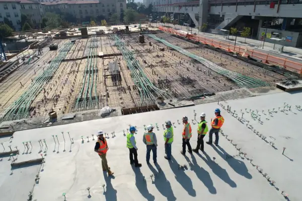 Grupa budowlańców w kaskach i kamizelkach obserwuje postęp prac na budowie, gdzie przygotowywana jest płyta fundamentowa zgodnie z art. 62 prawa budowlanego.