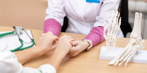 Arzt hält Patientin die Hand. Modelle von Fuß und Hand liegen auf dem Tisch. Hoffnung auf eine Wunderwaffe gegen Arthrose.
