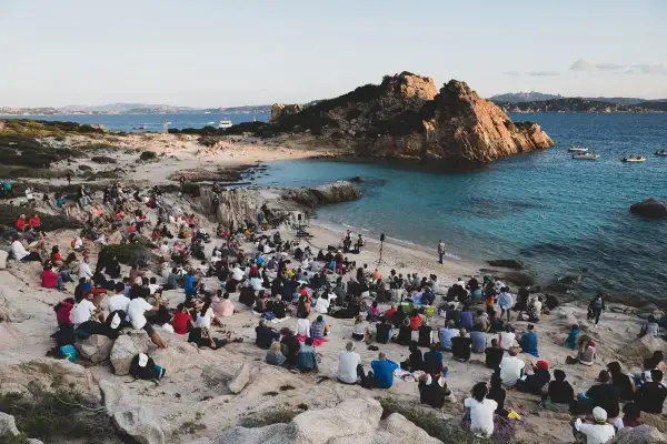 Concerto sulla spiaggia in Sardegna, con un pubblico seduto tra le rocce che ascolta un cantante sardo.