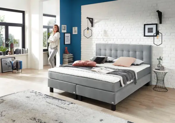 Modernes Boxspringbett in Grau, ideal für erholsamen Schlaf bei Bandscheibenvorfall. Eine Frau lehnt lässig daneben.