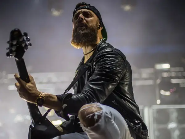 Charyzmatyczny gitarzysta z brodą podczas koncertu Bullet for My Valentine, w skórzanej kurtce i podartych spodniach, gra na gitarze.