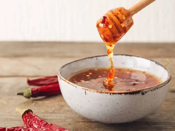 En träsked droppar gyllene chili honung ner i en skål. Torkade chilifrukter ligger bredvid.