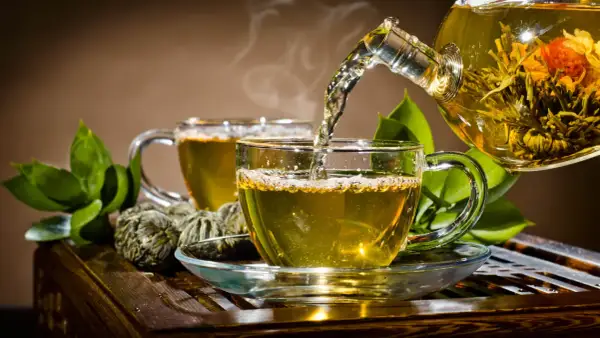 Gorąca herbata parzy się w szklanej filiżance, a z dzbanka wlewa się aromatyczny napar. Obok leżą zielone kulki herbaty.
