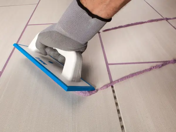 Mano con guanto grigio stende cemento e acqua senza sabbia con spatola su fughe di piastrelle.
