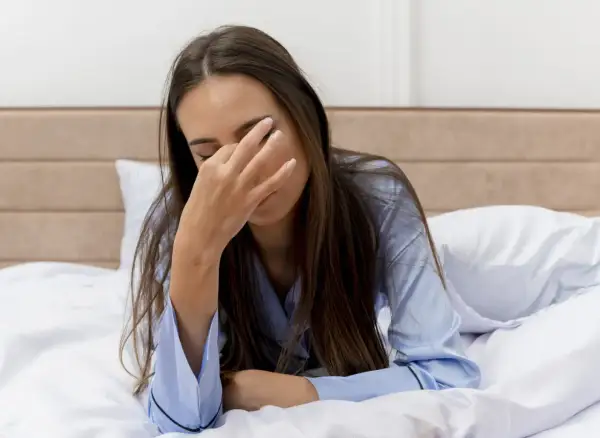 Een vrouw ligt moe in bed, haar hand voor haar ogen. Ze draagt een blauwe pyjama.