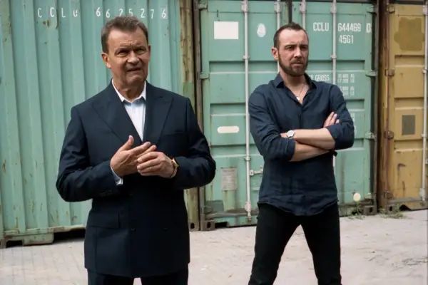 Dwóch mężczyzn, jeden w garniturze, drugi w koszuli, stoi przed kontenerami. Scena jak z filmu "Jak zostałem gangsterem. Cytaty" o życiu w półświatku.