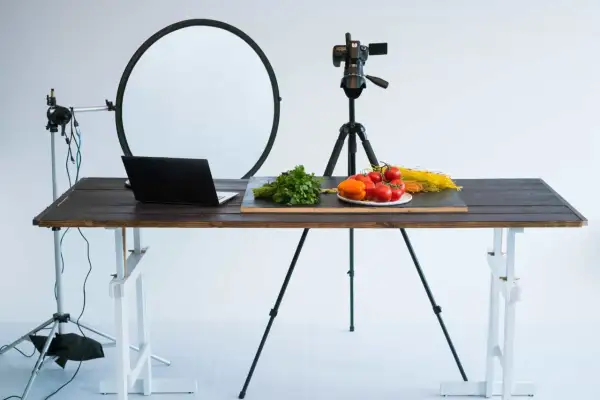 A tabletop photography setup with a camera on a tripod, a laptop, and fresh ingredients like tomatoes and pasta on a wooden table.
