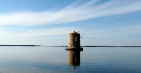 Mulino solitario nella laguna di Orbetello, riflesso sull'acqua calma sotto un cielo azzurro con nuvole sottili.