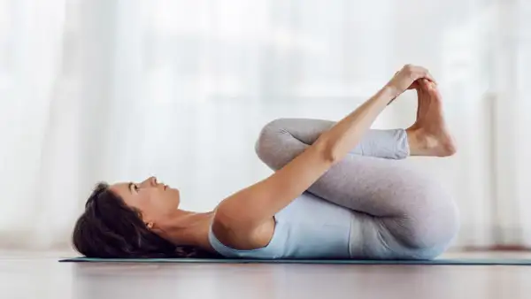 Mujer en postura del bebé feliz, abrazando sus pies con las manos, relajada en una esterilla de yoga.