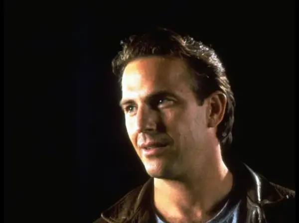 Kevin Costner jung, mit nachdenklichem Blick und brauner Lederjacke vor dunklem Hintergrund.