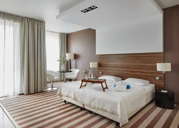 Przytulny pokój hotelowy z dwoma łóżkami, stolikiem i fotelami, ozdobiony drewnianym zagłówkiem i pasiastym dywanem.
