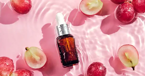 Odkryj naturalne olej z pestek winogron właściwości. Serum w buteleczce z pipetą otoczone świeżymi winogronami na różowym tle z falami wody.
