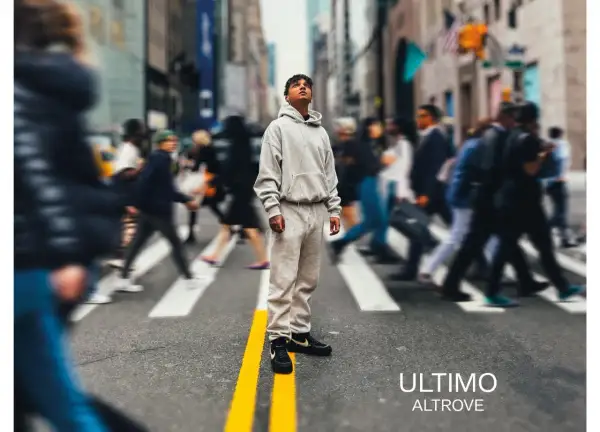 A young man in a grey tracksuit stands on a crosswalk in a busy city street, looking up.