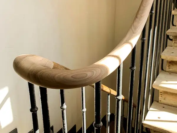 Courbe élégante d'une main courante en bois sur une volée d'escalier aux barreaux de fer forgé, baignée de lumière.