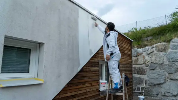 Mann streicht Fassade ohne Reinigung mit Roller. Ein Eimer Farbe steht bereit.