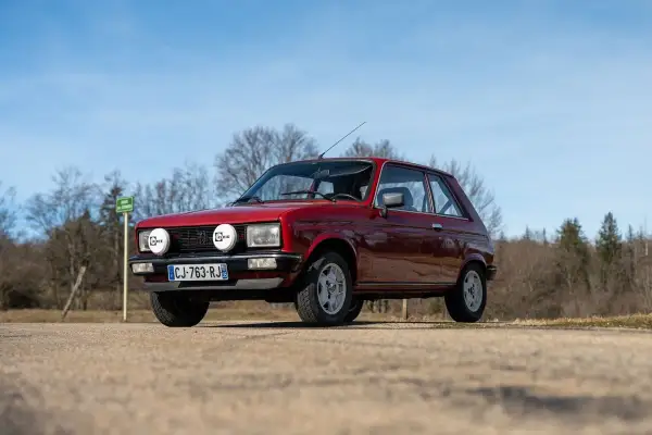 Röd Peugeot 104 med extraljus parkerad på en grusväg, med träd och blå himmel i bakgrunden.