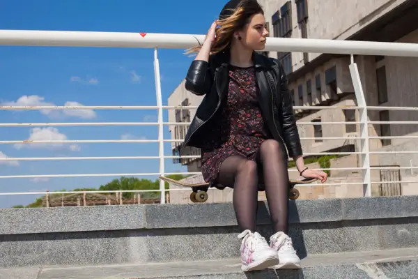 Dziewczyna w skórzanej kurtce i kwiecistej sukience, siedząca na deskorolce, prezentuje modne stylizacje z butami sportowymi.