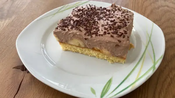Ein Stück Bananen Torte mit Schokoladencreme und Schokostreuseln auf einem Teller mit Blattmuster.